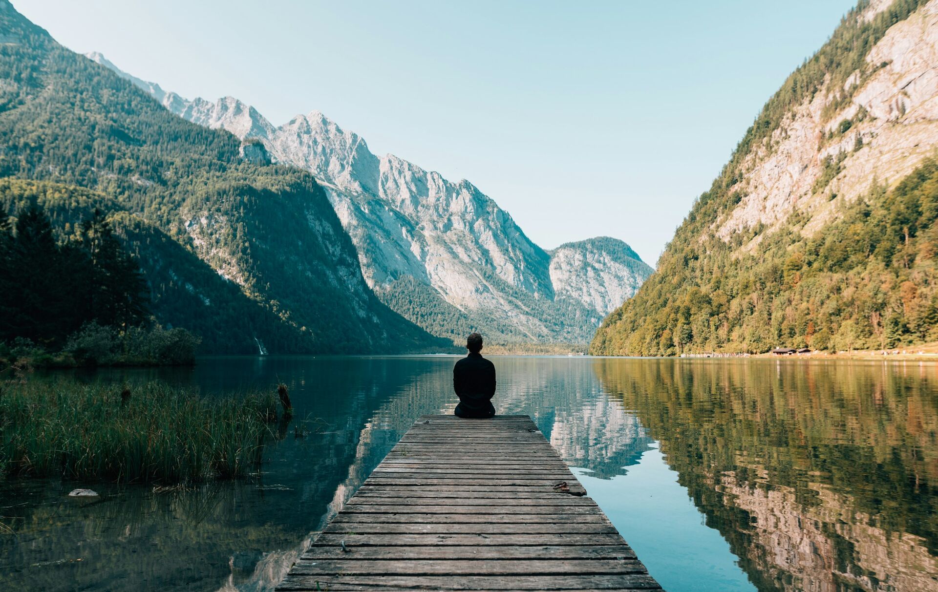 A figure sitting alone at the end of a jetty on a still mountain lake